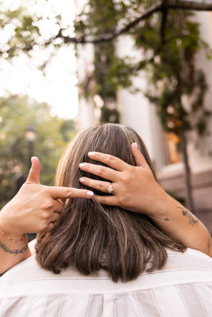 A playful portrait after an LGBTQ proposal in Savannah shows one woman embracing her partner from behind while spreading both hands across her hair, prominently displaying the sparkling engagement ring on her left hand while pointing to it with her right