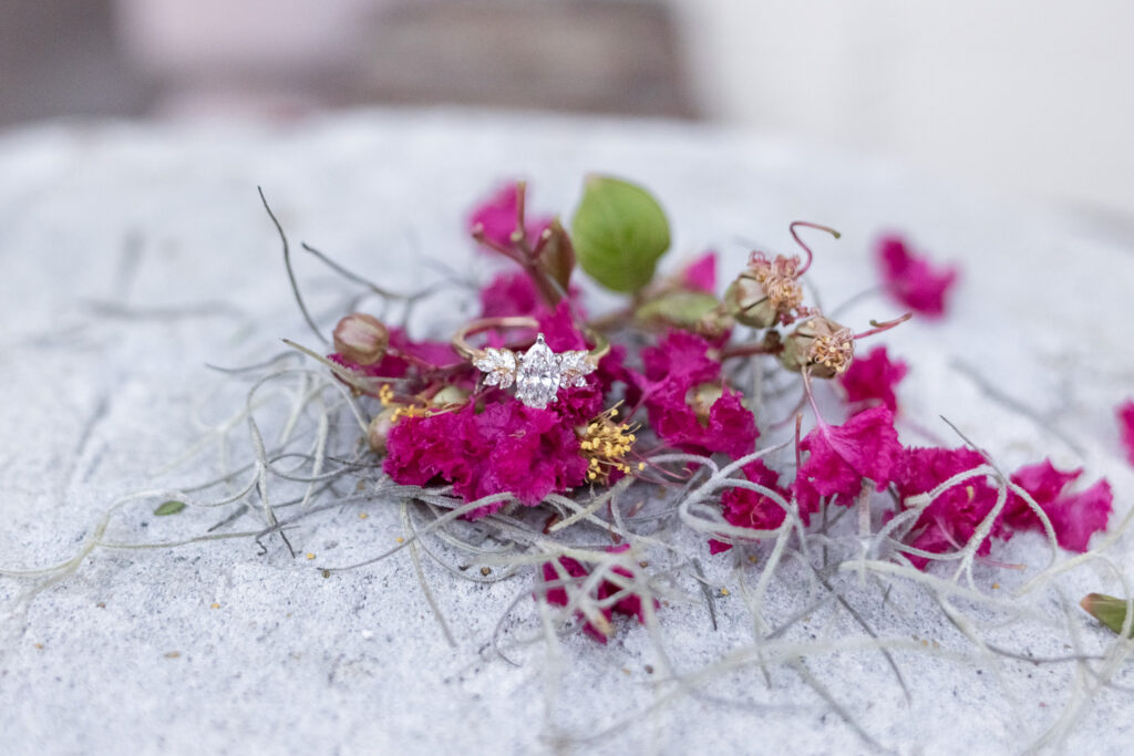 A Savannah proposal photographer creates a close detail portrait of the diamond engagement ring nestled among vivid magenta flowers, dried seed pods and wispy strands of gray Spanish moss on a pale stone surface.