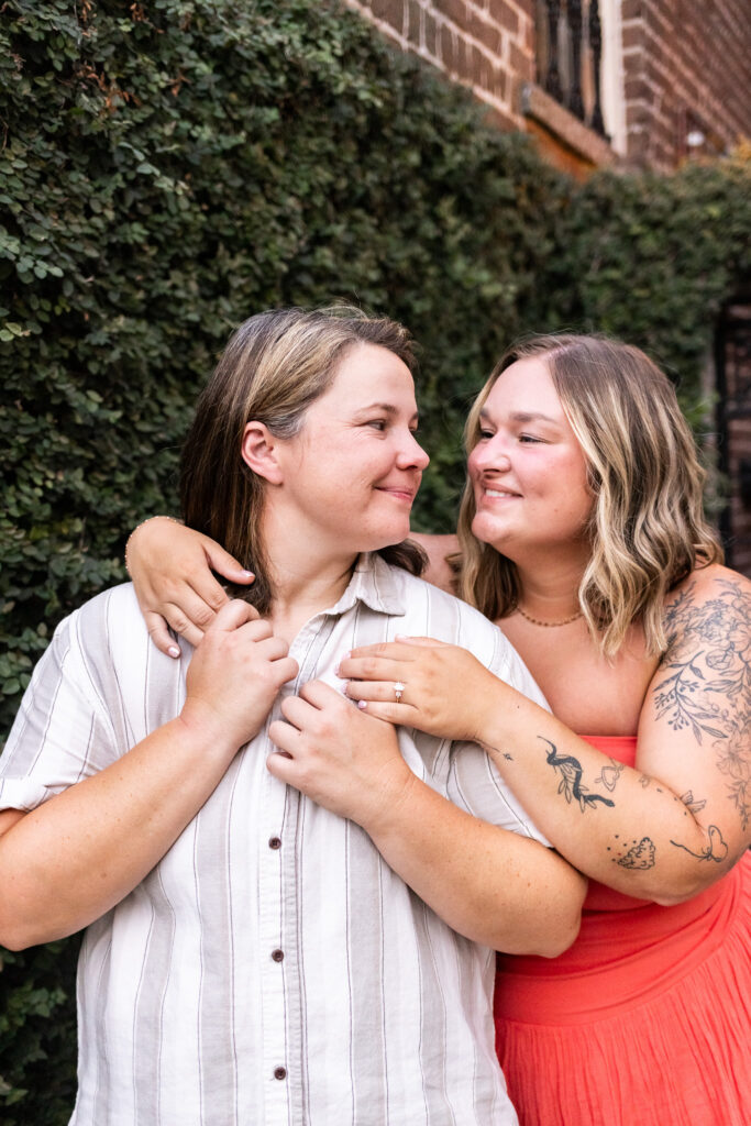 During portraits with their Savannah proposal photographer, one newly engaged woman wraps her arm around her partner and kisses her cheek in front of dense green ivy and an ornate black iron gate, with her new ring resting against her partner’s chest.