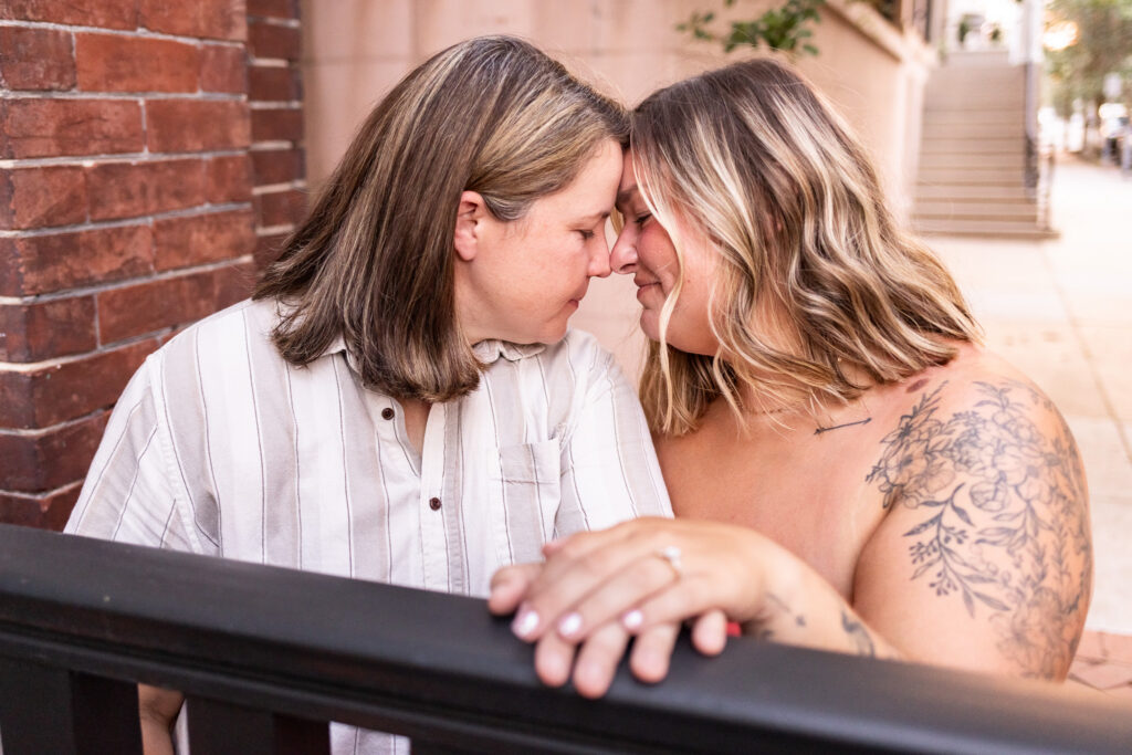 Two newly engaged women lean across a black railing until their foreheads and noses touch, both closing their eyes while one rests her ring hand prominently in the foreground.