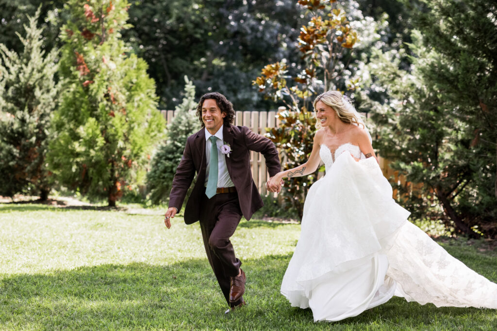 A newlywed couple holds hands and runs together across a sunlit lawn surrounded by lush trees during their South Jersey wedding portraits.
