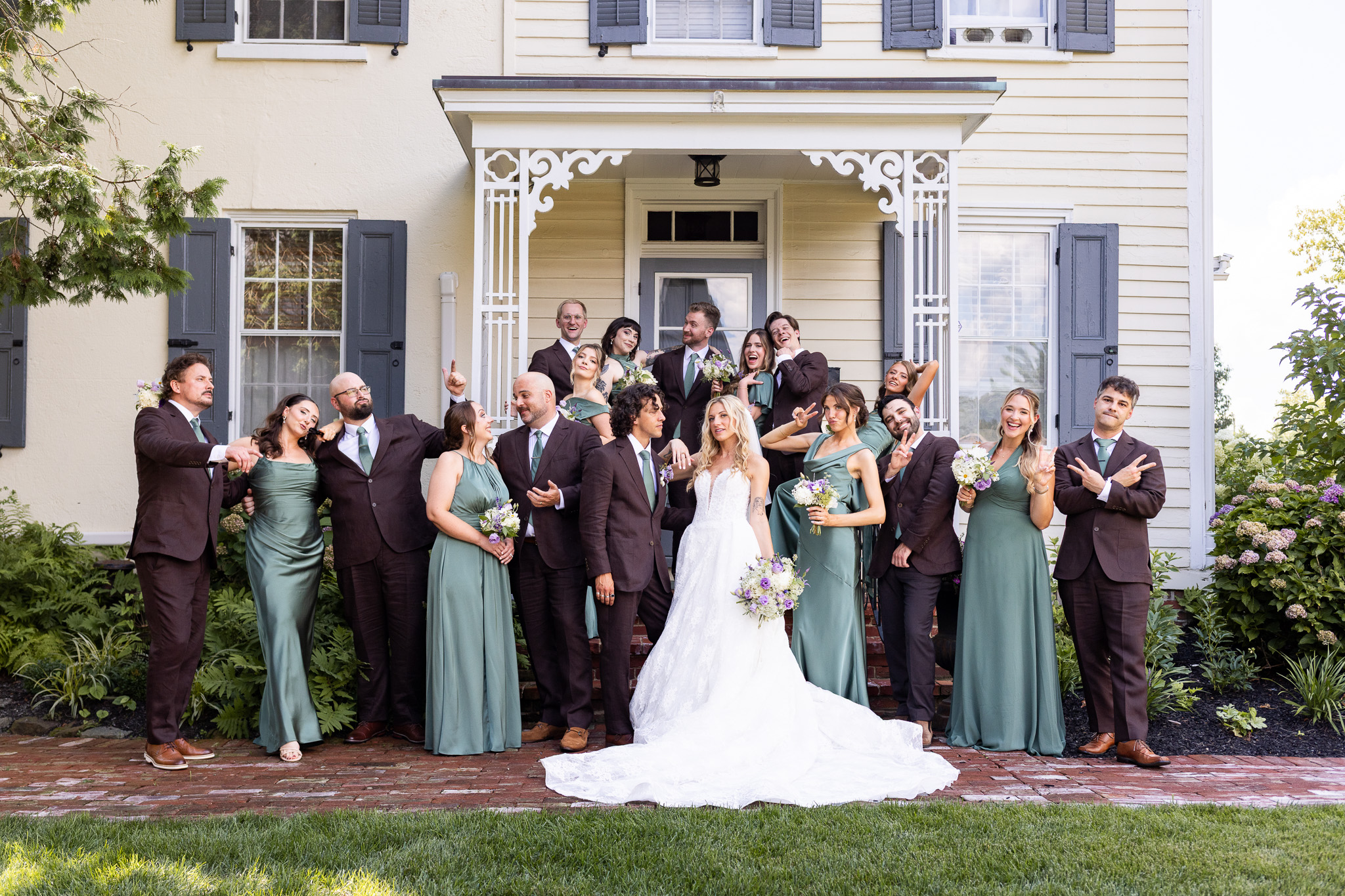 A newlywed couple stands at the center of their wedding party as everyone laughs, poses and gestures playfully in front of the historic farmhouse.