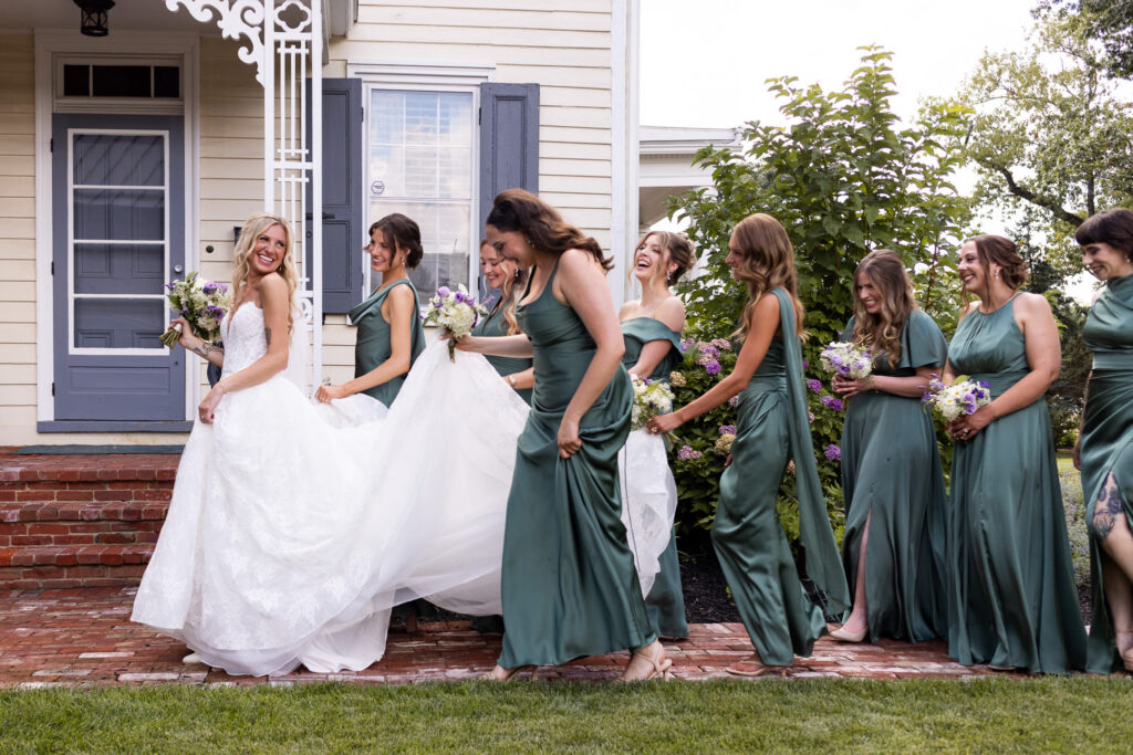 A bride laughs with her wedding party in sage green dresses as they walk beside a historic farmhouse during a joyful South Jersey wedding.