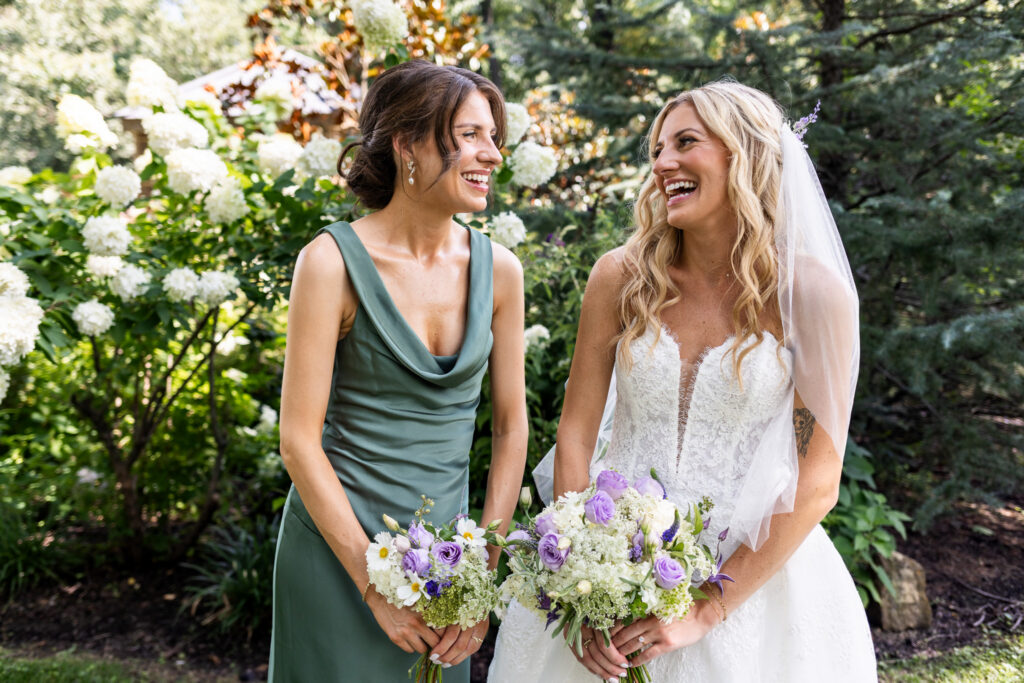 A bride and bridesmaid laugh together while holding purple and white bouquets in a garden filled with white hydrangeas and greenery.
