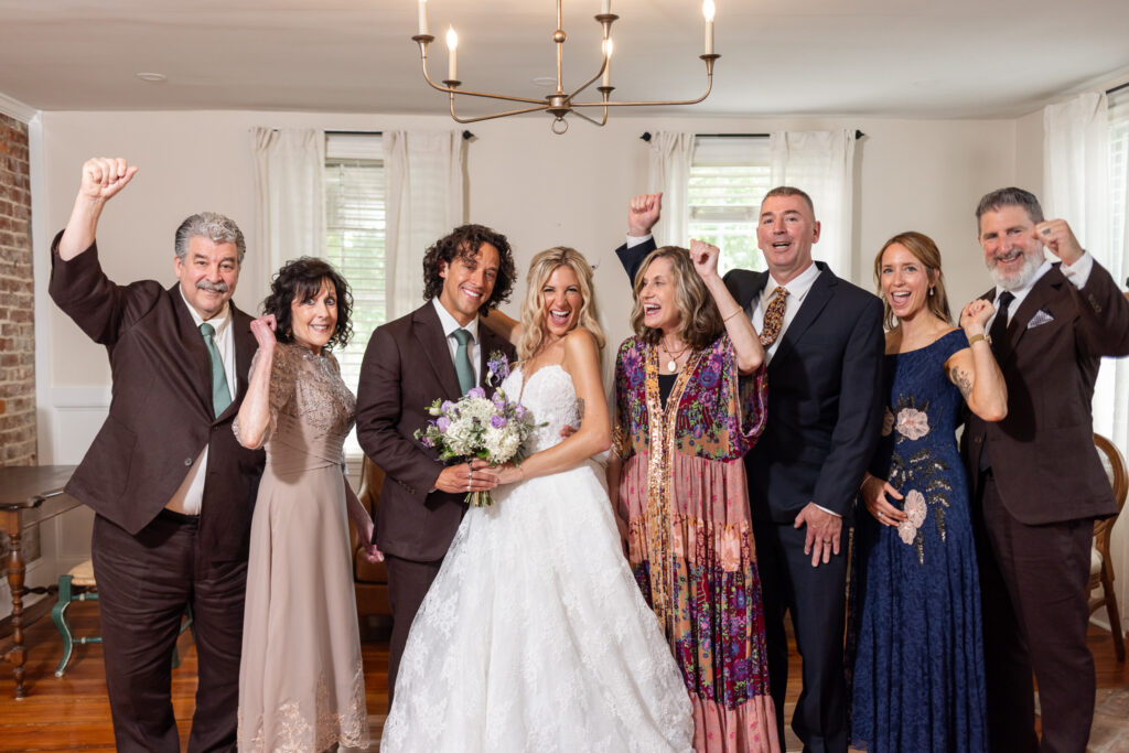 A bride and groom stand surrounded by family members inside a Magnolia, NJ wedding venue, everyone laughing and raising their fists in celebration beneath a simple metal chandelier.