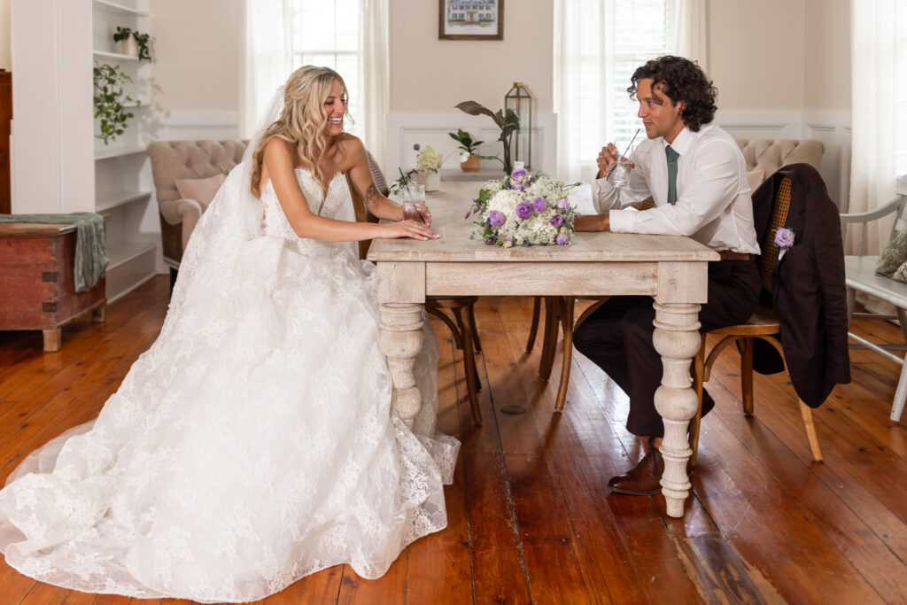 A newlywed couple relaxes across from one another at a rustic wooden table inside a Magnolia, NJ wedding venue, smiling over glasses of water with the bride’s white and lavender bouquet arranged between them.
