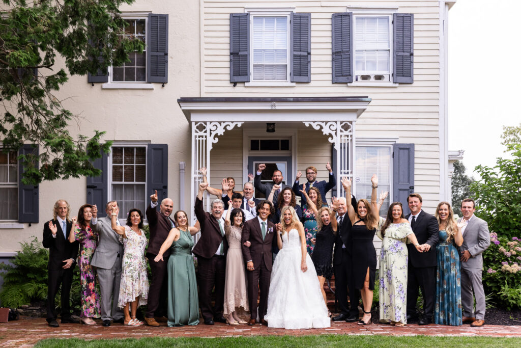 A large group of family members celebrates outside the historic farmhouse during a Founders Grove wedding, raising their hands while the newlyweds stand together in the center.