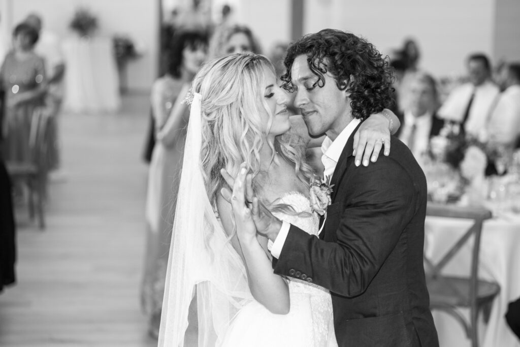 A black-and-white photograph captures the newlyweds dancing closely together during their reception as guests watch from the surrounding tables.