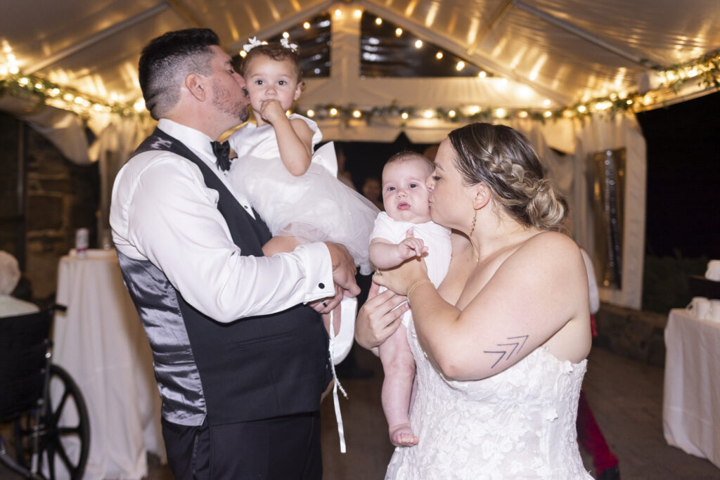 The newlyweds hold their two young children during the evening wedding reception beneath glowing string lights. The groom kisses the older child on the cheek while the bride kisses the infant she is holding, capturing an affectionate family moment amid the celebration.