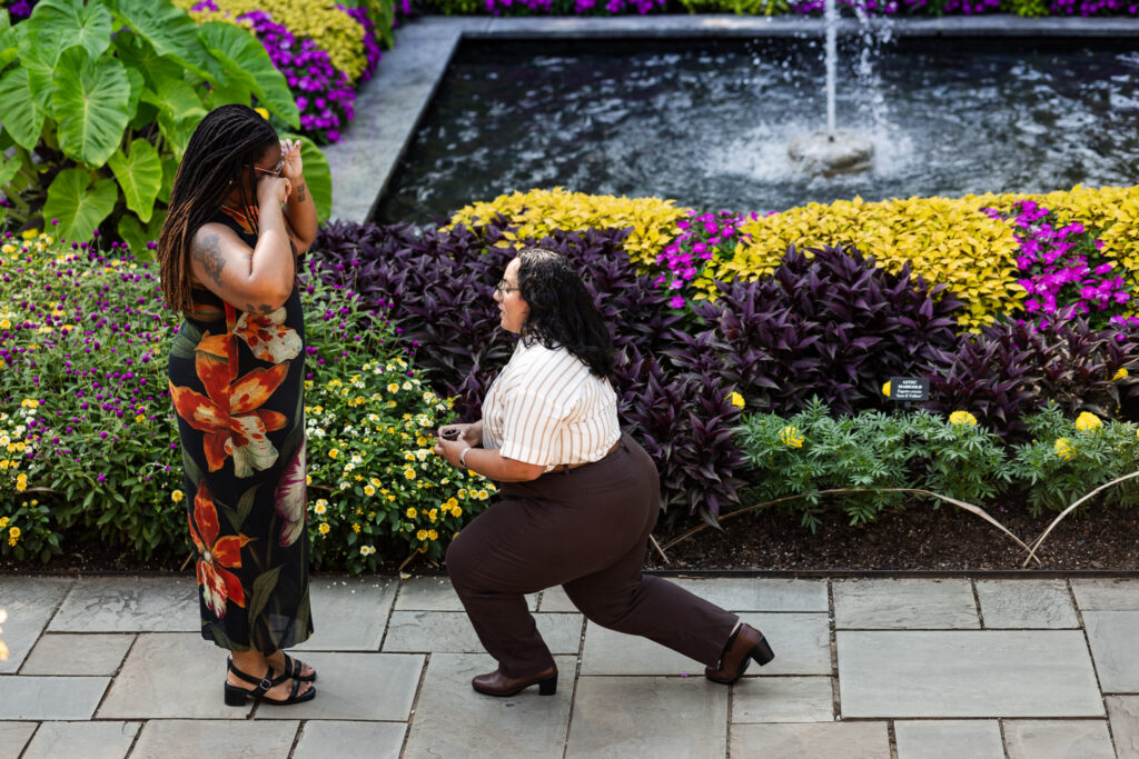 Longwood Gardens lesbian proposal beside a rectangular fountain surrounded by vivid purple, yellow and green foliage, as one partner kneels with the ring while the other covers her face in an emotional reaction.