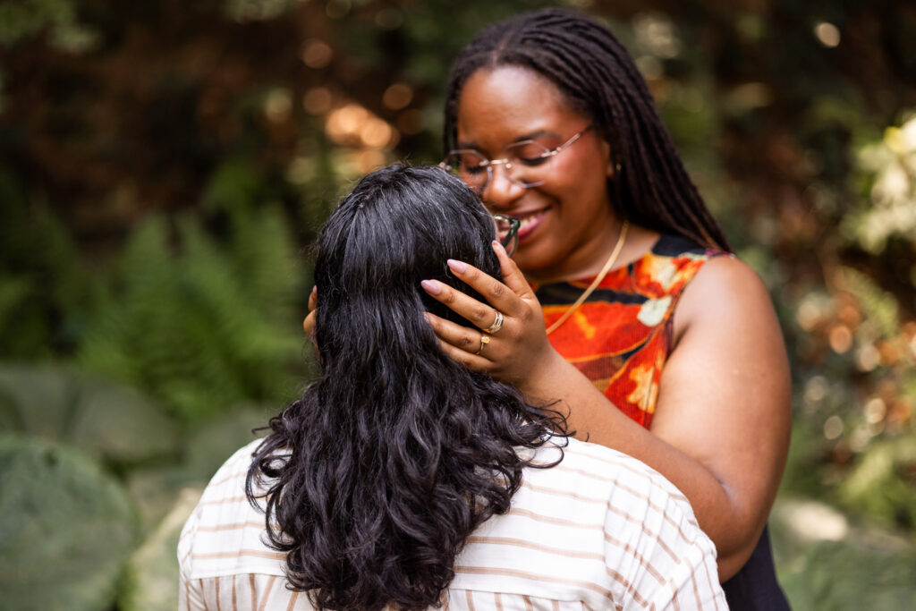 One partner gently holds her fiancée's face, smiling down at her beneath deep green trees in a softly shaded garden.