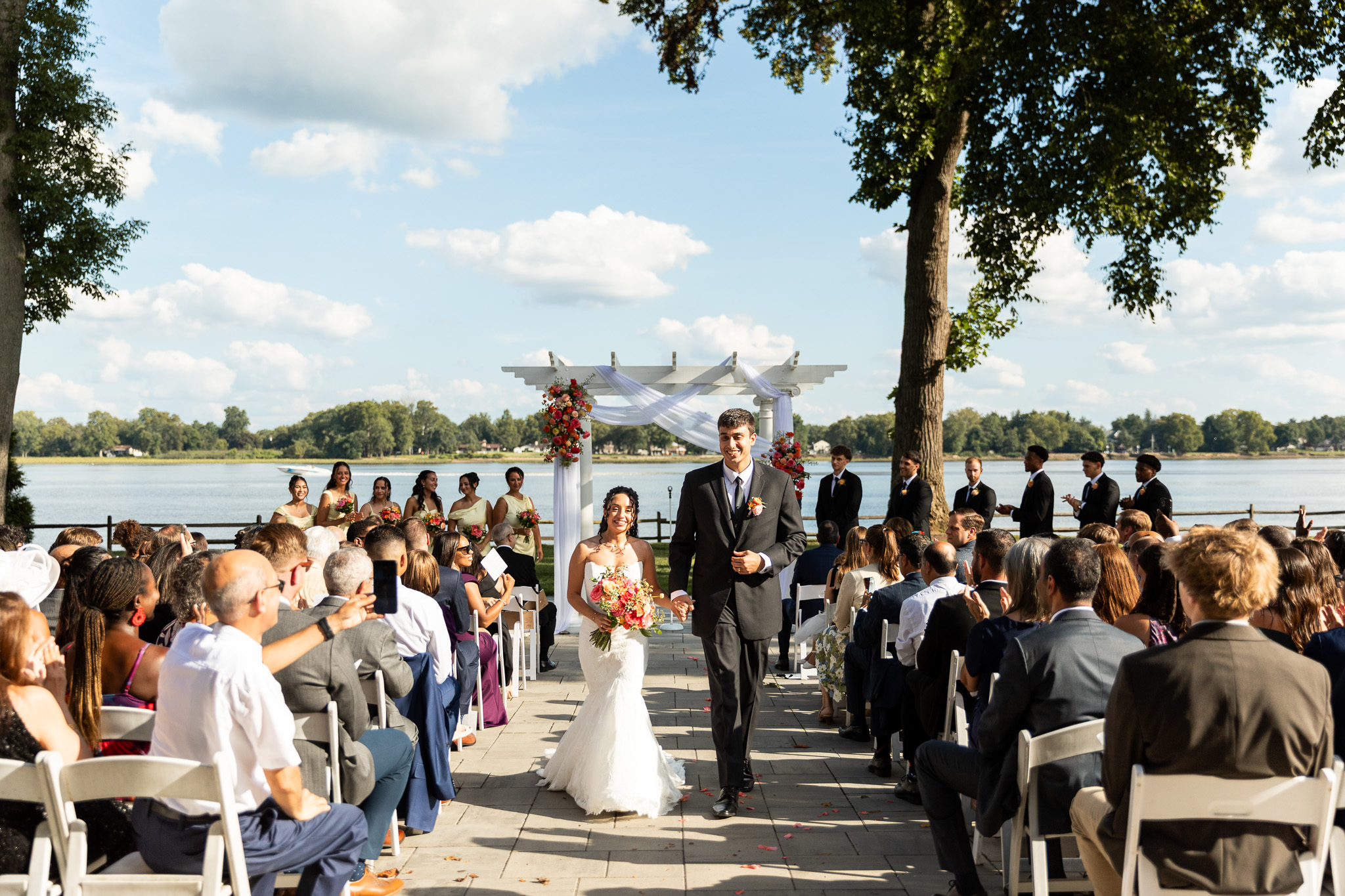 Bride and groom walk hand in hand back up the ceremony aisle as newlyweds while guests seated in white chairs applaud on both sides. Among the pen ryn rivers edge photos from their ceremony, the wide view shows the couple framed by their colorful flower-covered white arbor, wedding party and expansive Delaware River beneath a bright blue sky filled with soft clouds.
