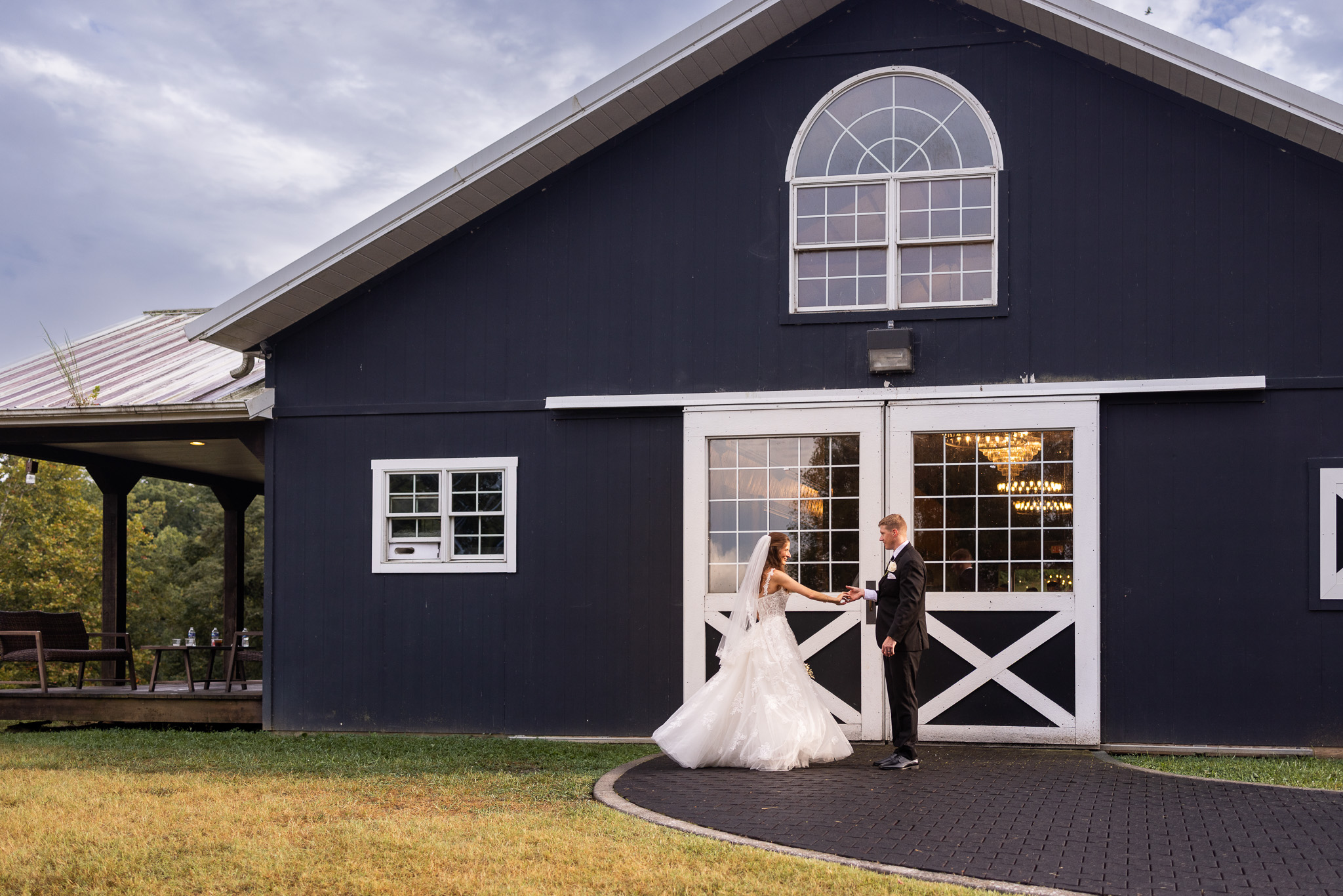 The groom twirls his bride in a wide shot in front of The Stables at Strawberry Creek. They appear small beneath its dramatic peaked roof and oversized arched window. Warm chandeliers glow through the white barn doors behind them while a moody, cloud-filled sky and surrounding lawn give the portrait a sweeping, romantic sense of place.