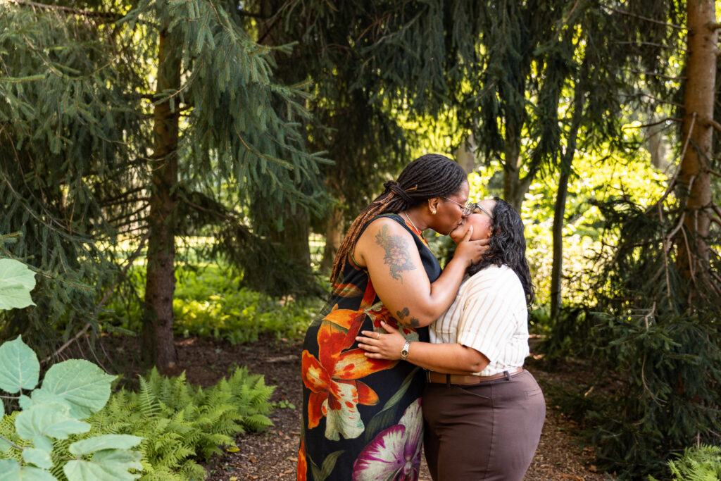 Couple kissing beneath towering evergreen branches during their Longwood Gardens lesbian proposal, framed by ferns, textured foliage and warm sunlight filtering through the trees.