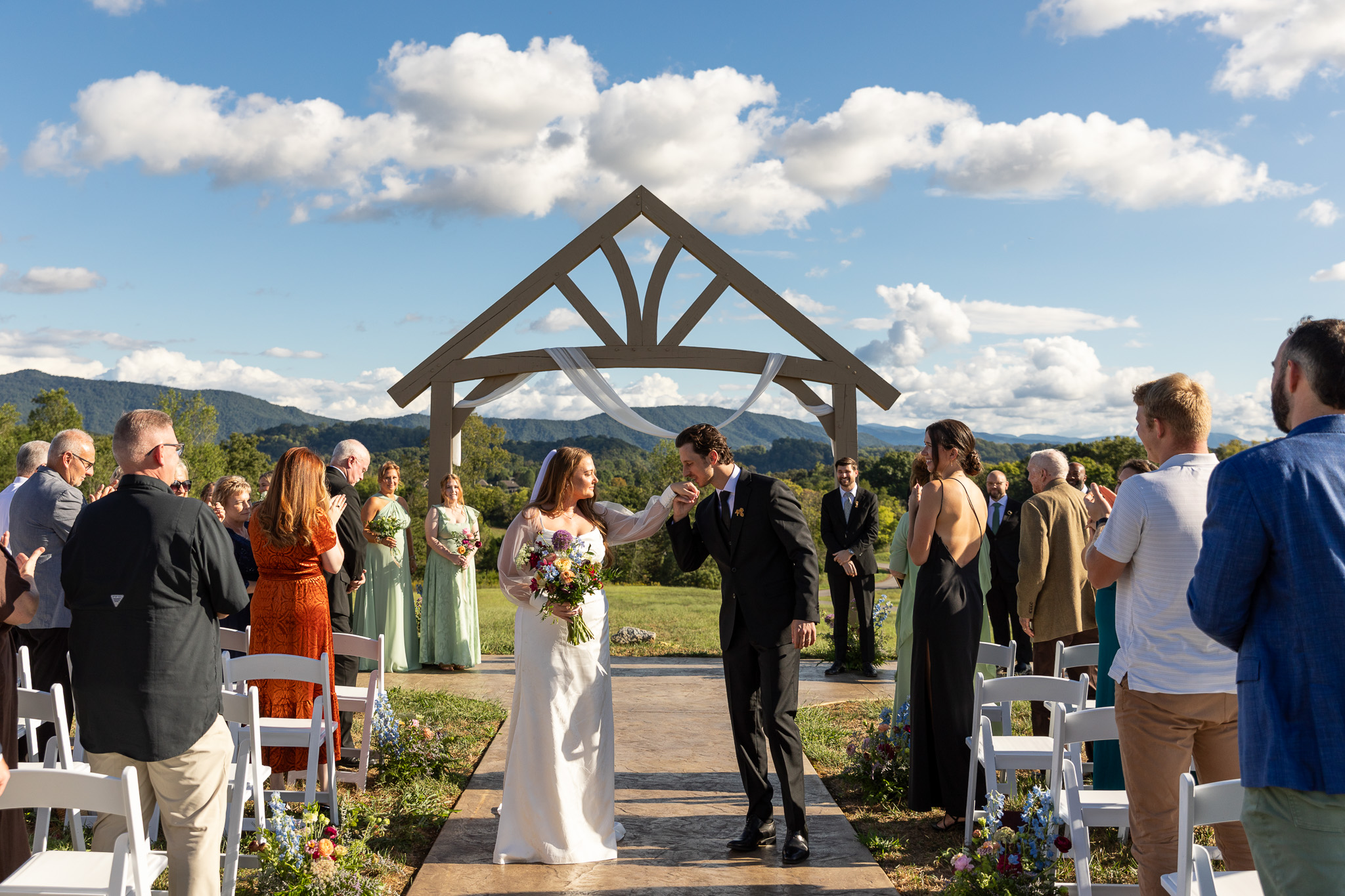 Newlyweds walk back down the outdoor Ayoka Ranch wedding ceremony aisle as the groom kisses his bride's hand, surrounded by applauding wedding guests seated in white chairs. A wooden peaked arbor draped in soft white fabric frames the distant mountain views, while colorful wildflower arrangements line the aisle beneath a bright blue sky.