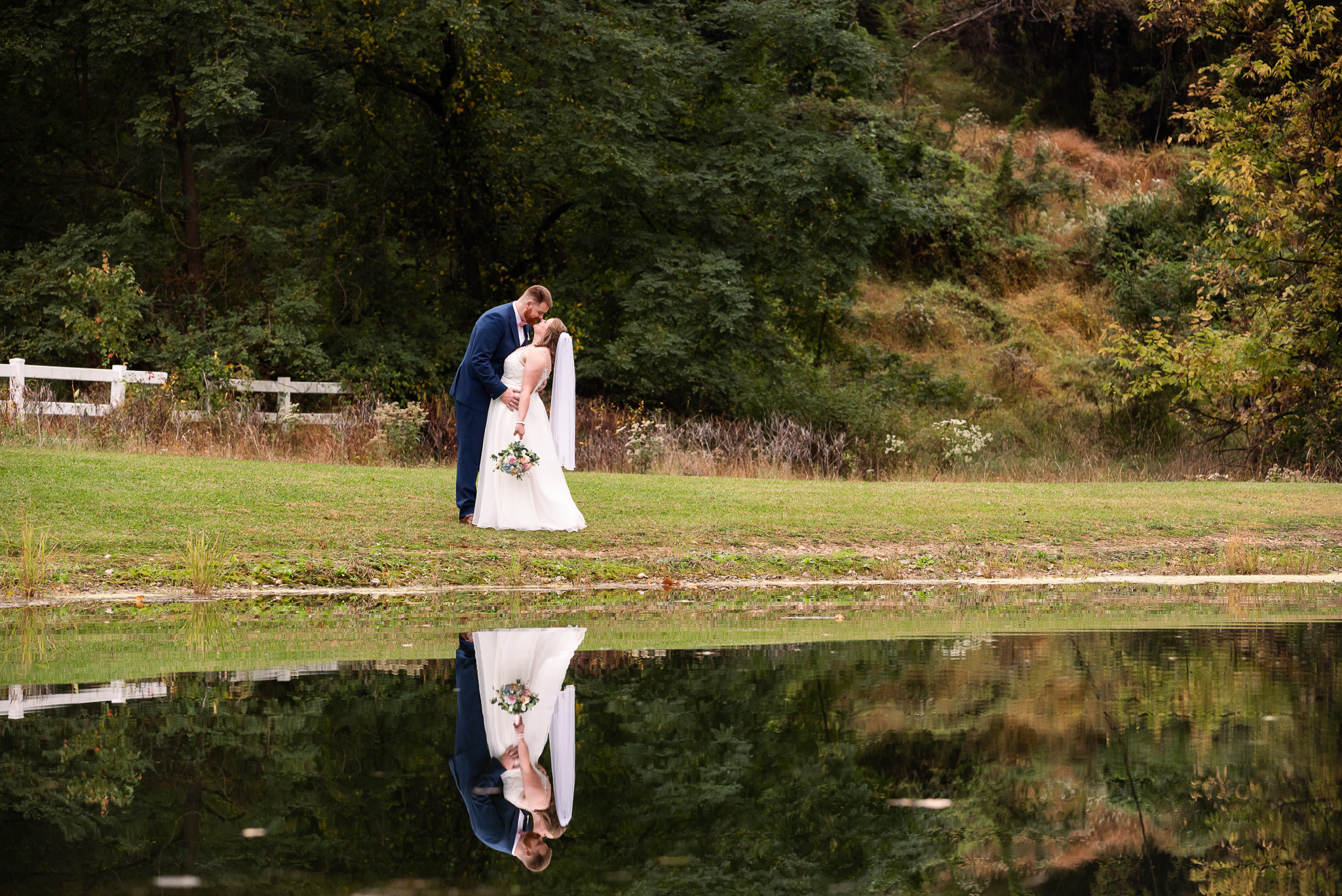 A bride and groom share a kiss beside a still pond at a Harrisburg wedding venue, their full-length reflections mirrored in the dark water. The bride's flowing white gown and veil contrast with the groom's navy suit, while dense green trees, wild grasses, and a rustic white fence create a peaceful early autumn backdrop.